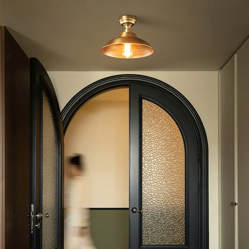 Warm glow inside the Antique Brass shade of this Clérachic ceiling light, hung over arched black doors and a passing figure.