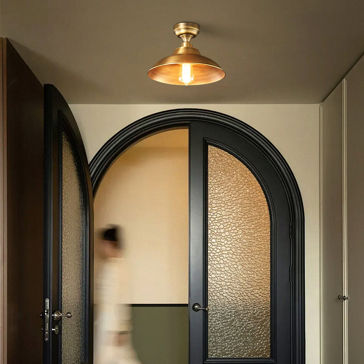 Warm glow inside the Antique Brass shade of this Clérachic ceiling light, hung over arched black doors and a passing figure.