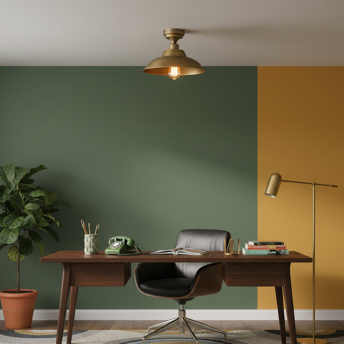 In a study with a green and mustard wall, a Clérachic brass dome ceiling light hangs over a walnut desk, a leather chair and a floor lamp.