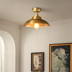 Close view of the Antique Brass dome and its warm filament bulb on this Clérachic ceiling light, with an arched window and a framed print behind.
