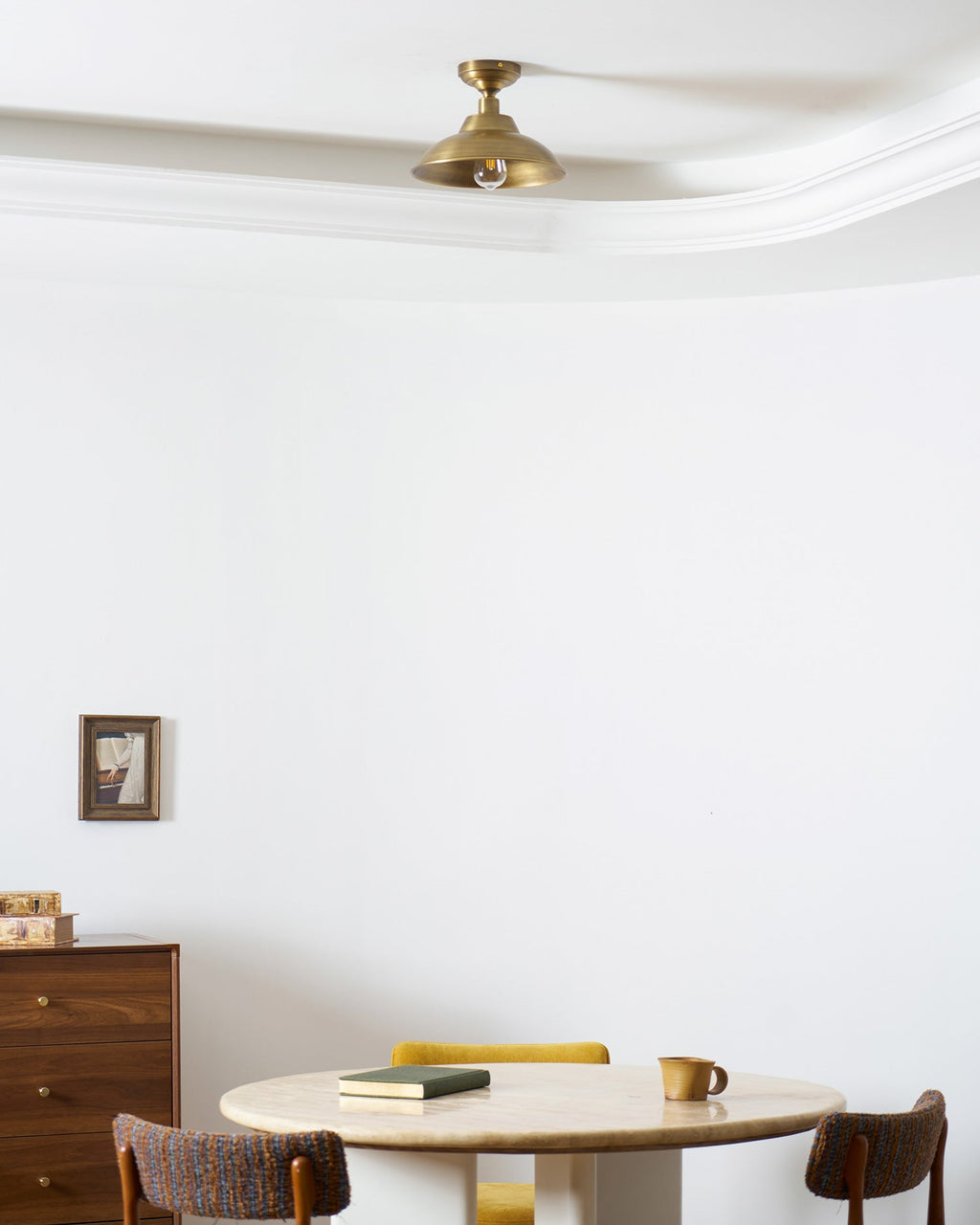 High on a white ceiling by a curved cornice, a Clérachic brass dome light lit warm, with a small framed picture and a dining table far below.