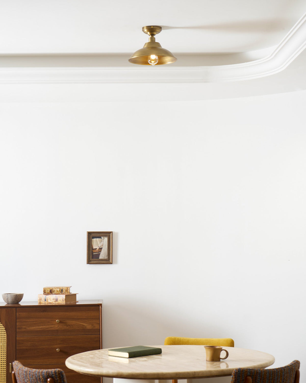 A small Clérachic brass dome ceiling light against a curved white cornice, with a round table and cord-seat chairs in the room beneath.