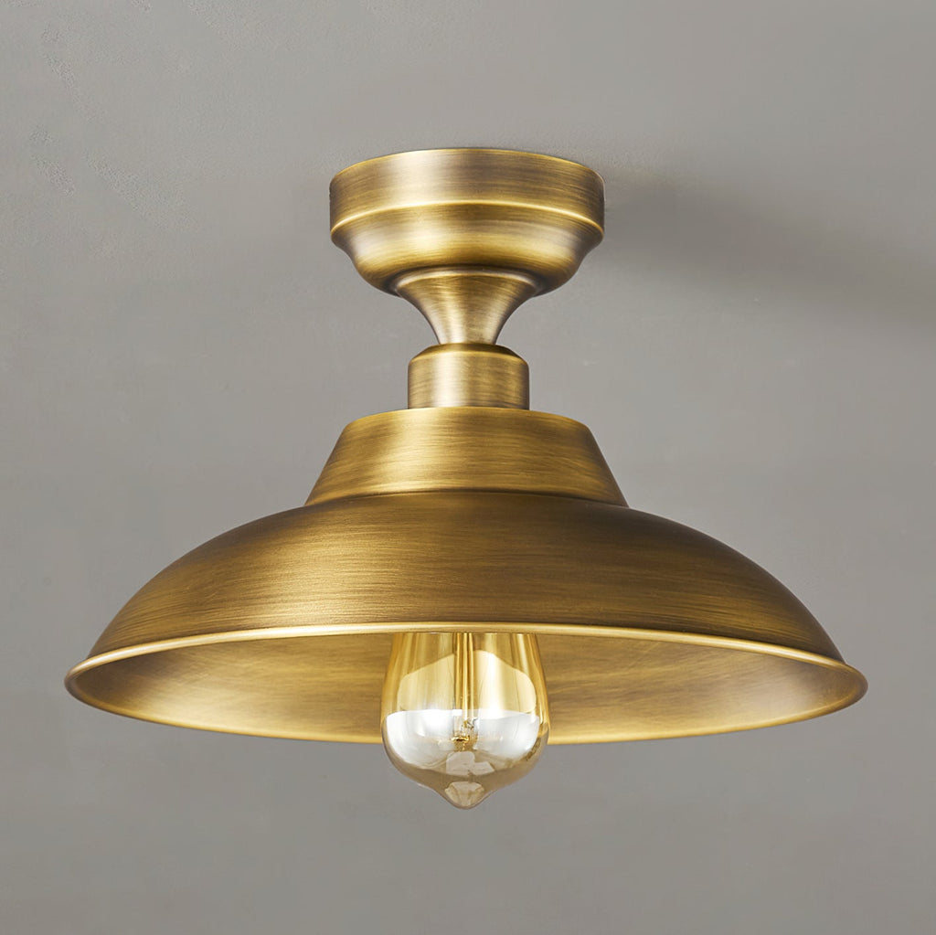Close view of this Clérachic ceiling light in Antique Brass, with a brushed finish on the dome and a clear bulb on a grey backdrop.