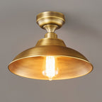 A Clérachic ceiling light with its brass dome lit from inside, the warm bulb glowing against a dark grey backdrop.