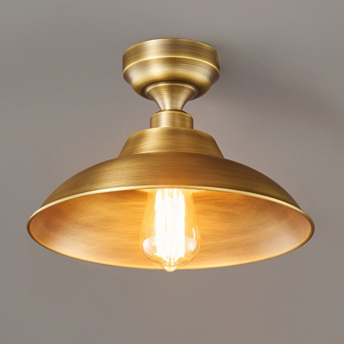A Clérachic ceiling light with its brass dome lit from inside, the warm bulb glowing against a dark grey backdrop.