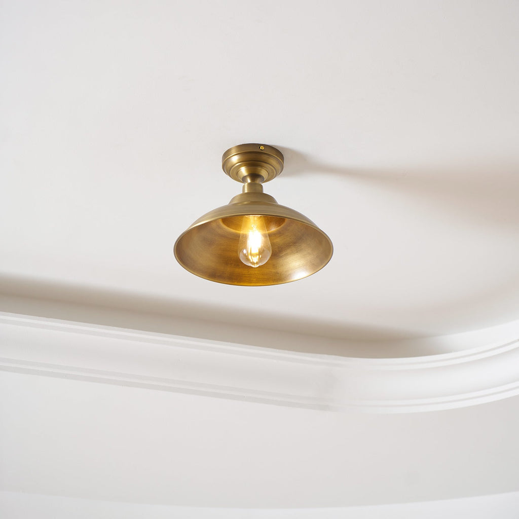 Fitted flush to a white ceiling beside a cornice, this Clérachic brass dome light glows with a warm bulb inside the shade.