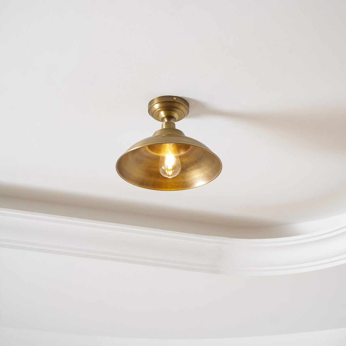 Fitted flush to a white ceiling beside a cornice, this Clérachic brass dome light glows with a warm bulb inside the shade.