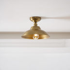 A Clérachic brass dome ceiling light fitted tight to a white ceiling above a plain wall, its warm bulb lit inside the dome.