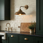 Lit above a kitchen counter, a Clérachic brass wall light hangs in front of cream tiles beside dark green cabinets and a brass tap.