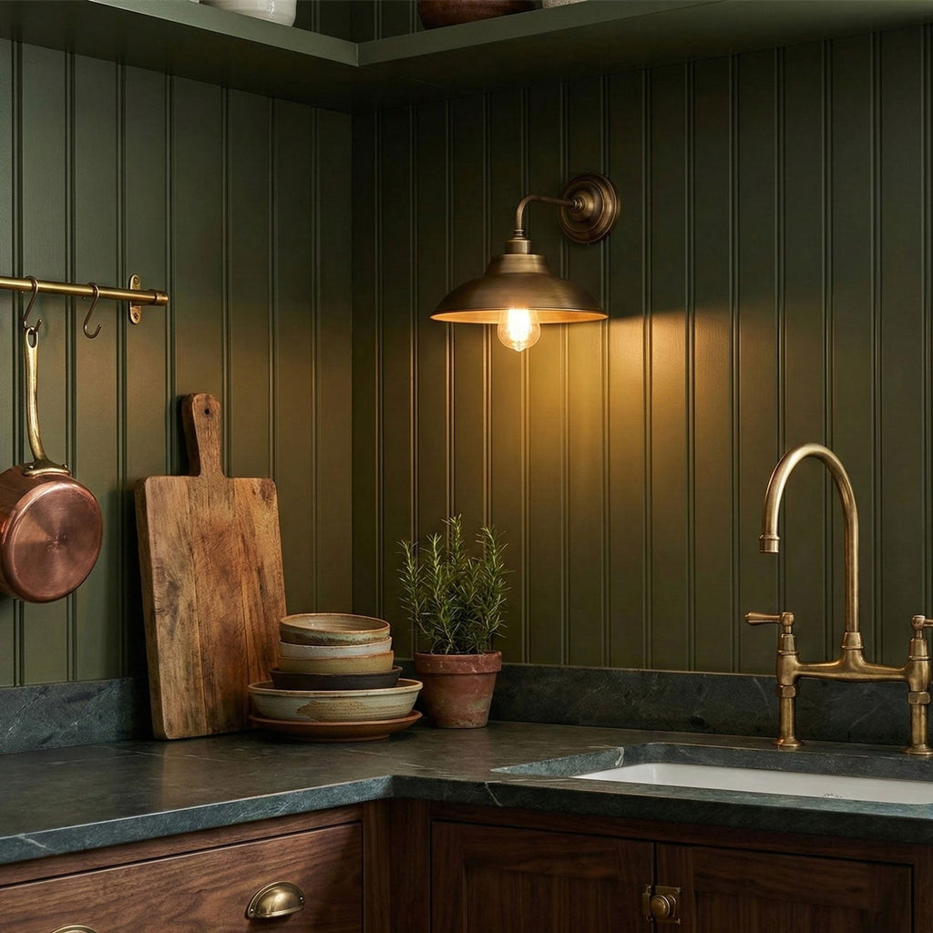 On green panelled kitchen walls, this Clérachic antique brass wall light shines warmly above a stone worktop and a brass tap.