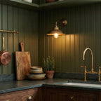 On green panelled kitchen walls, this Clérachic antique brass wall light shines warmly above a stone worktop and a brass tap.