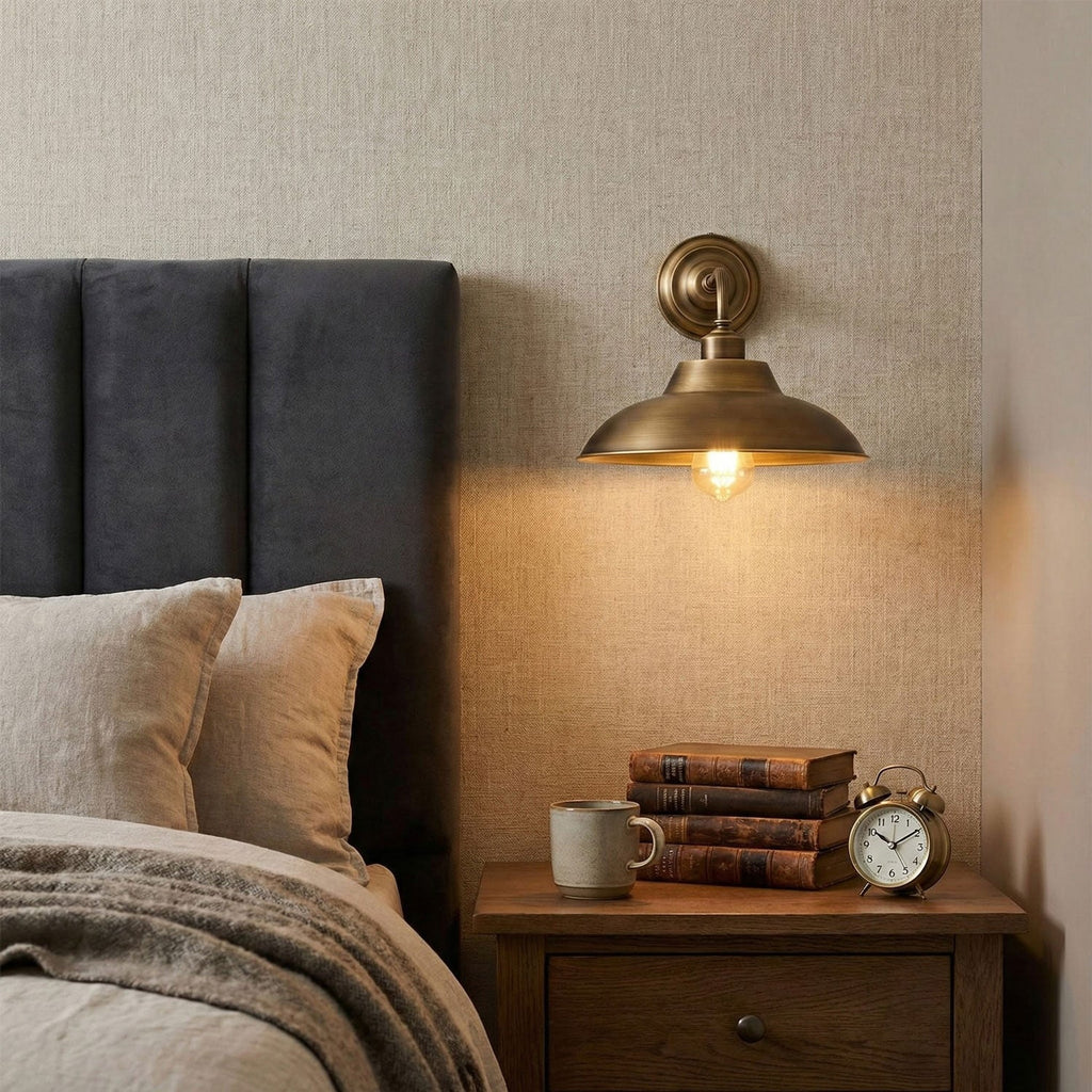 Beside a grey upholstered headboard, a Clérachic brass wall light casts a warm pool of light over a bedside table with books and a clock.