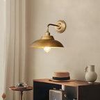 A Clérachic antique brass wall light on a warm beige wall, with a straw hat and a small table with a cup beneath it.