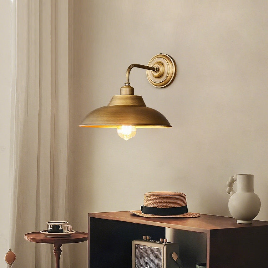 A Clérachic antique brass wall light on a warm beige wall, with a straw hat and a small table with a cup beneath it.
