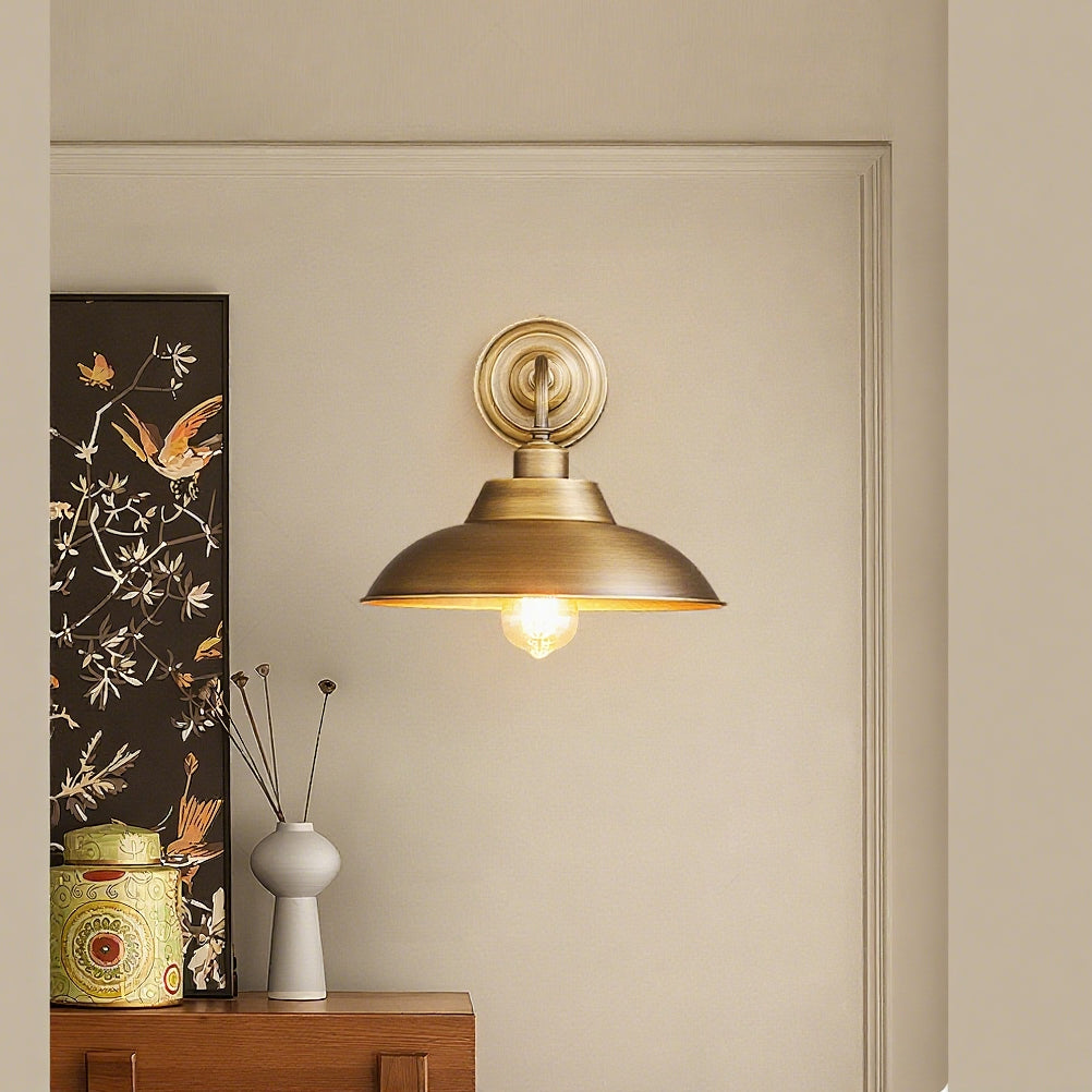 A Clérachic brass wall light hangs on a pale wall beside a dark floral panel, above a wooden unit with a tin and a vase.