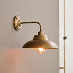 Seen from the side, this Clérachic antique brass wall light shows its round backplate, curved arm and dome shade with a warm bulb.