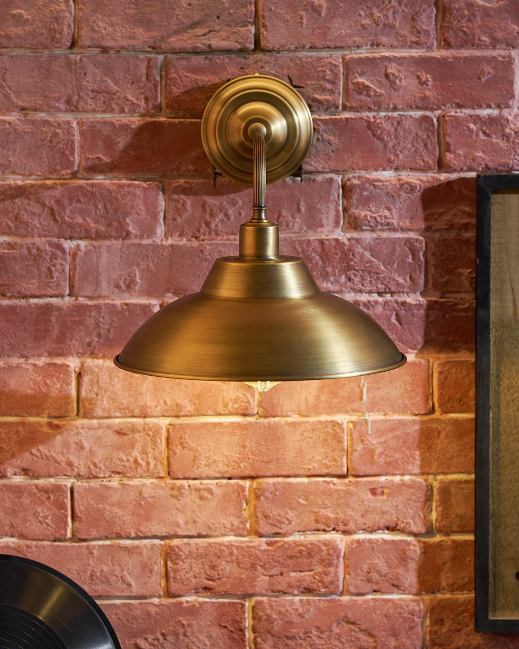 A Clérachic brass wall light casts a warm glow down a red brick wall, with the dome shade seen from slightly below.