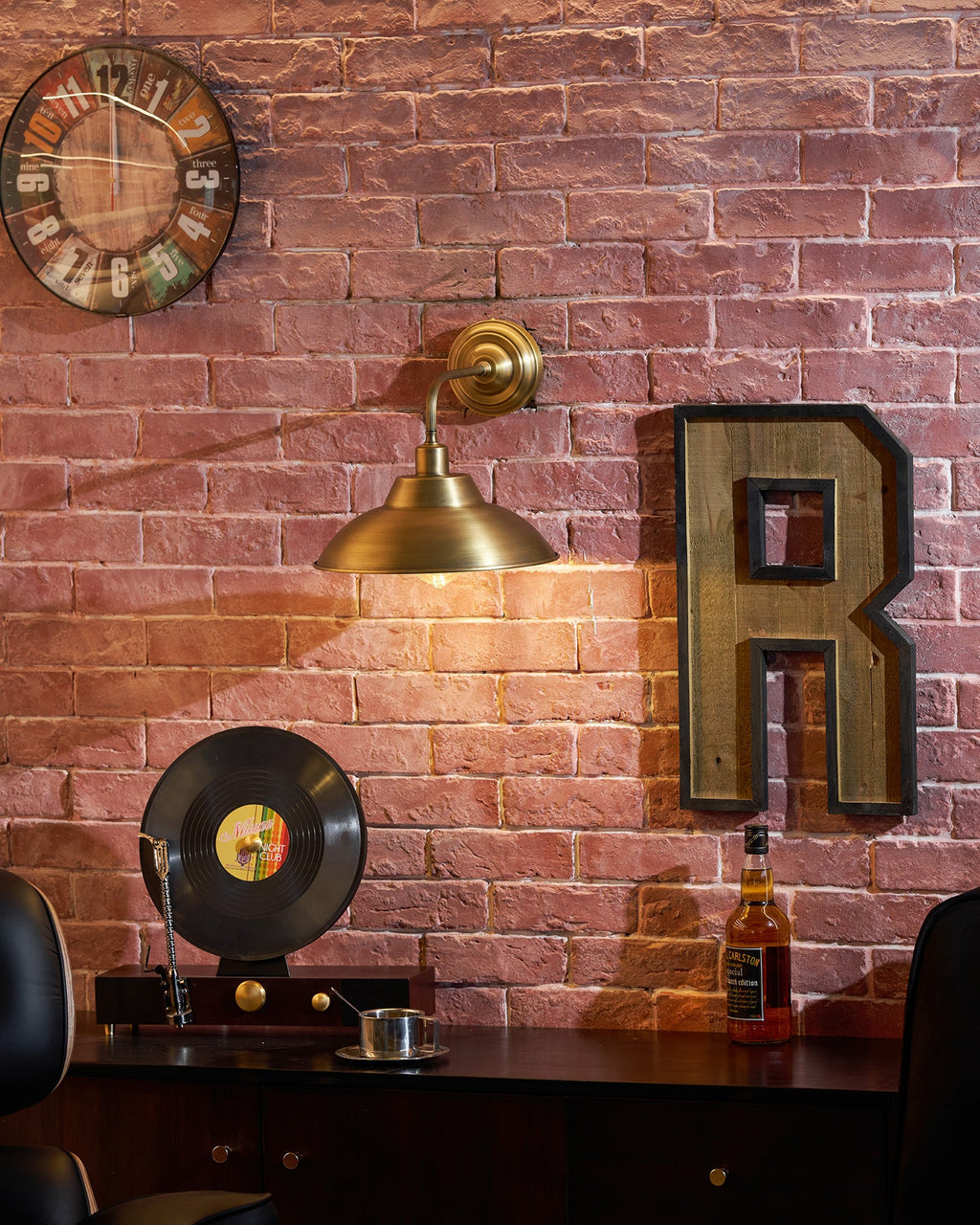 A Clérachic brass wall light on a brick wall above a dark sideboard, with a large wooden letter R, a wall clock and a record player.