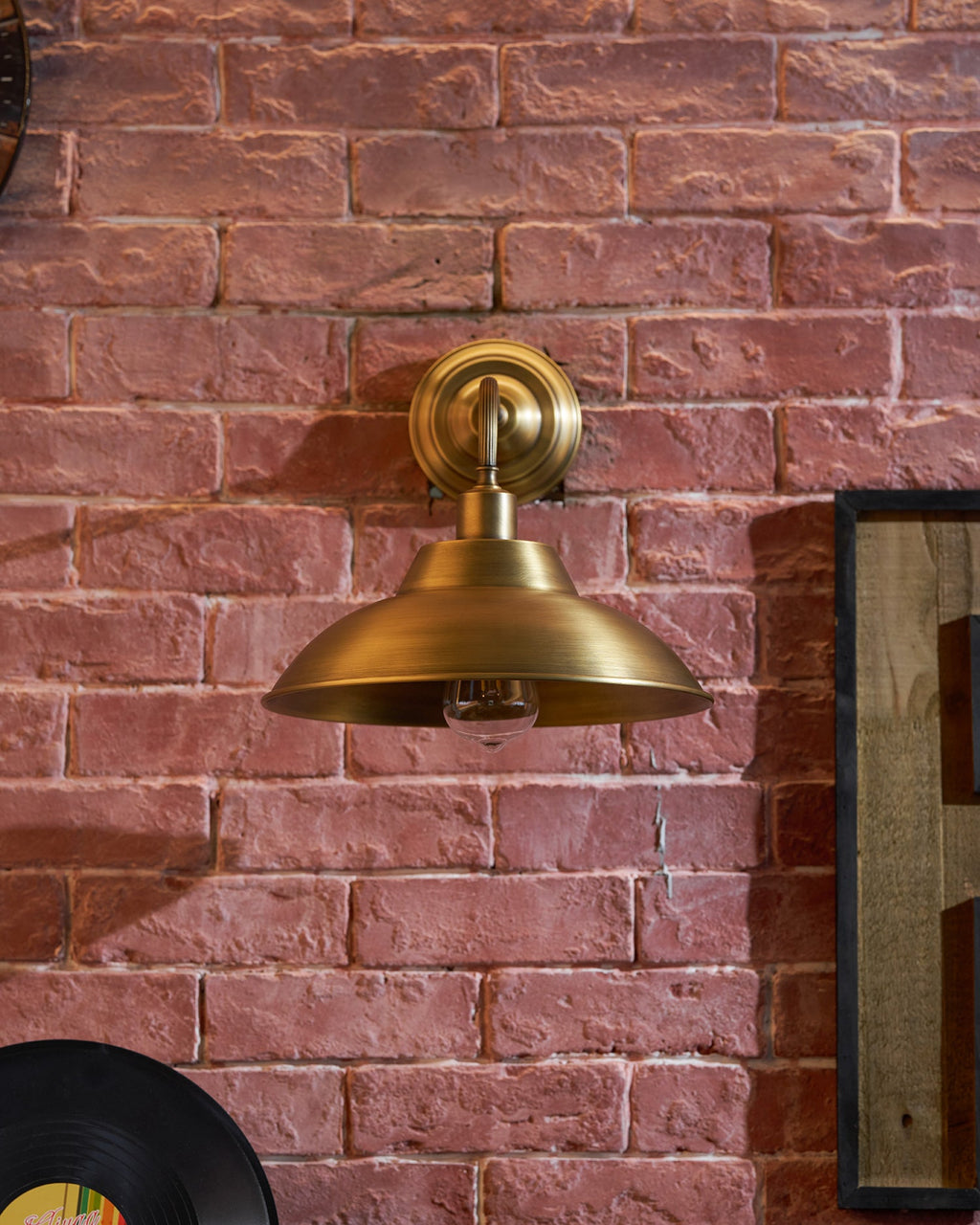 This Clérachic antique brass wall light hangs on red brick, with the edge of a vinyl record and a wooden sign beside it.