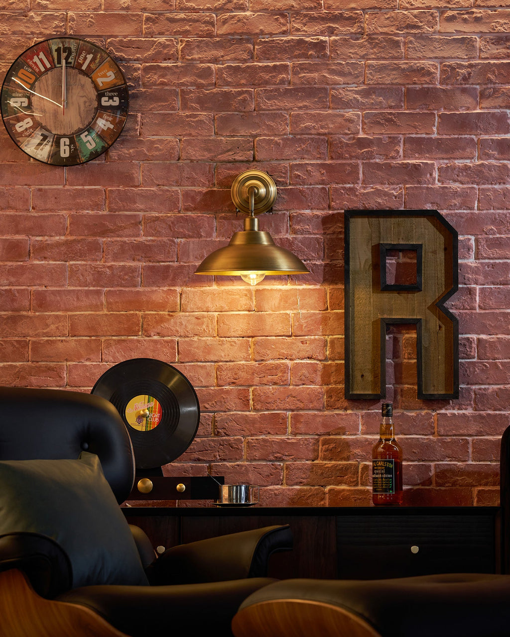 A Clérachic brass wall light lights a brick wall above a black leather armchair, with a wall clock, a letter R sign and a record.