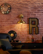 A Clérachic brass wall light lights a brick wall above a black leather armchair, with a wall clock, a letter R sign and a record.