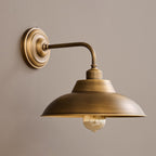 Close three-quarter view of this Clérachic antique brass wall light on a taupe wall, with its curved arm and clear bulb below the shade.