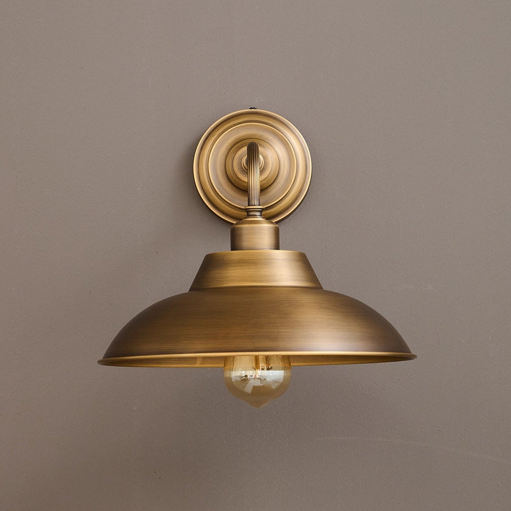 Straight-on view of this Clérachic brass wall light on a taupe wall, showing the round backplate and wide dome shade.