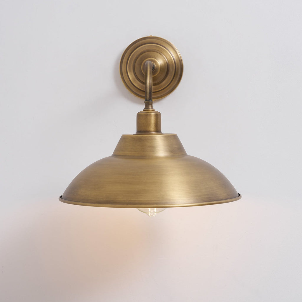 This Clérachic brass wall light on a pale grey wall throws a soft warm glow beneath its dome shade.
