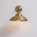 A lit Clérachic antique brass wall light on a pale wall, with the warm bulb showing under the shade and a glow on the wall below.