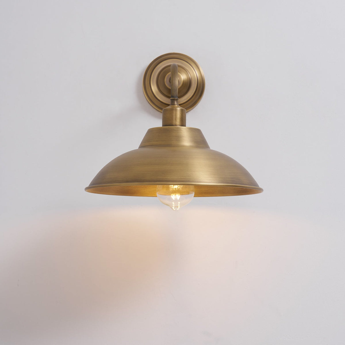A lit Clérachic antique brass wall light on a pale wall, with the warm bulb showing under the shade and a glow on the wall below.