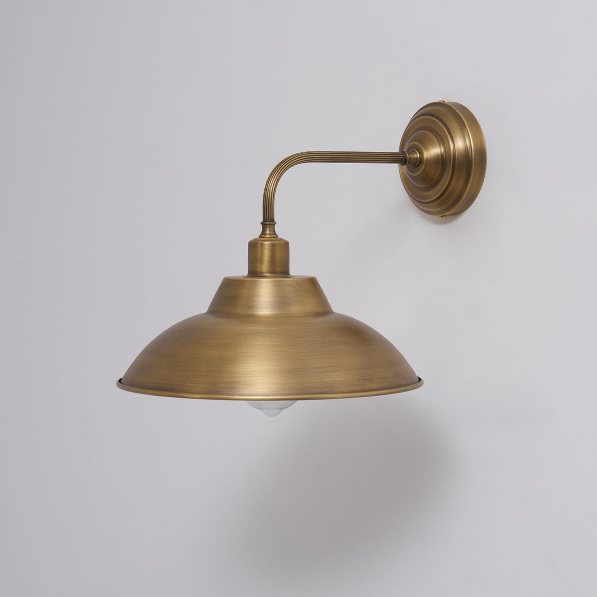 Side view of this Clérachic brass wall light on a pale grey wall, with the curved arm reaching out from a stepped round backplate.