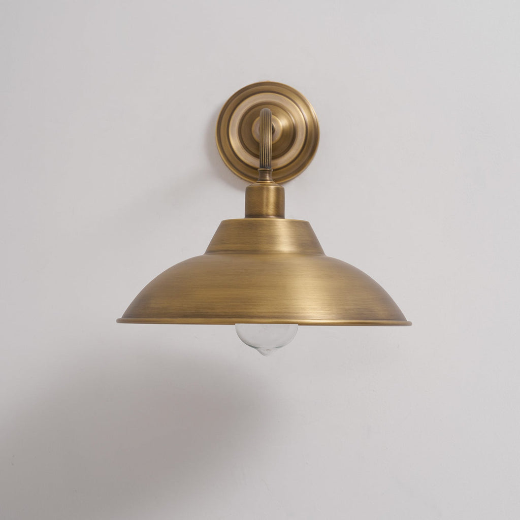 This Clérachic brass wall light, unlit, on a pale grey wall, showing the stepped round backplate and the dome shade with a clear bulb.