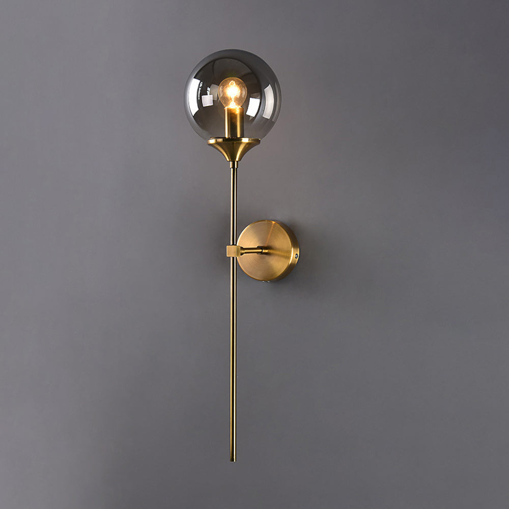 Clérachic Brass-Finish Globe Wall Sconce in smoke gray glass, lit against a soft grey wall with the brass stem running past the round plate.
