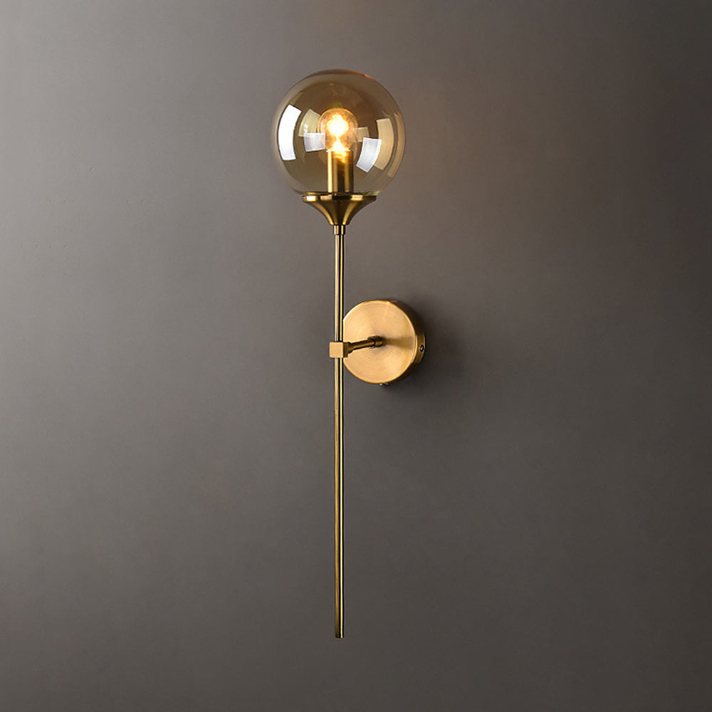 The amber glass option of the Clérachic Brass-Finish Globe Wall Sconce, glowing honey above its brushed brass stem on a warm grey wall.