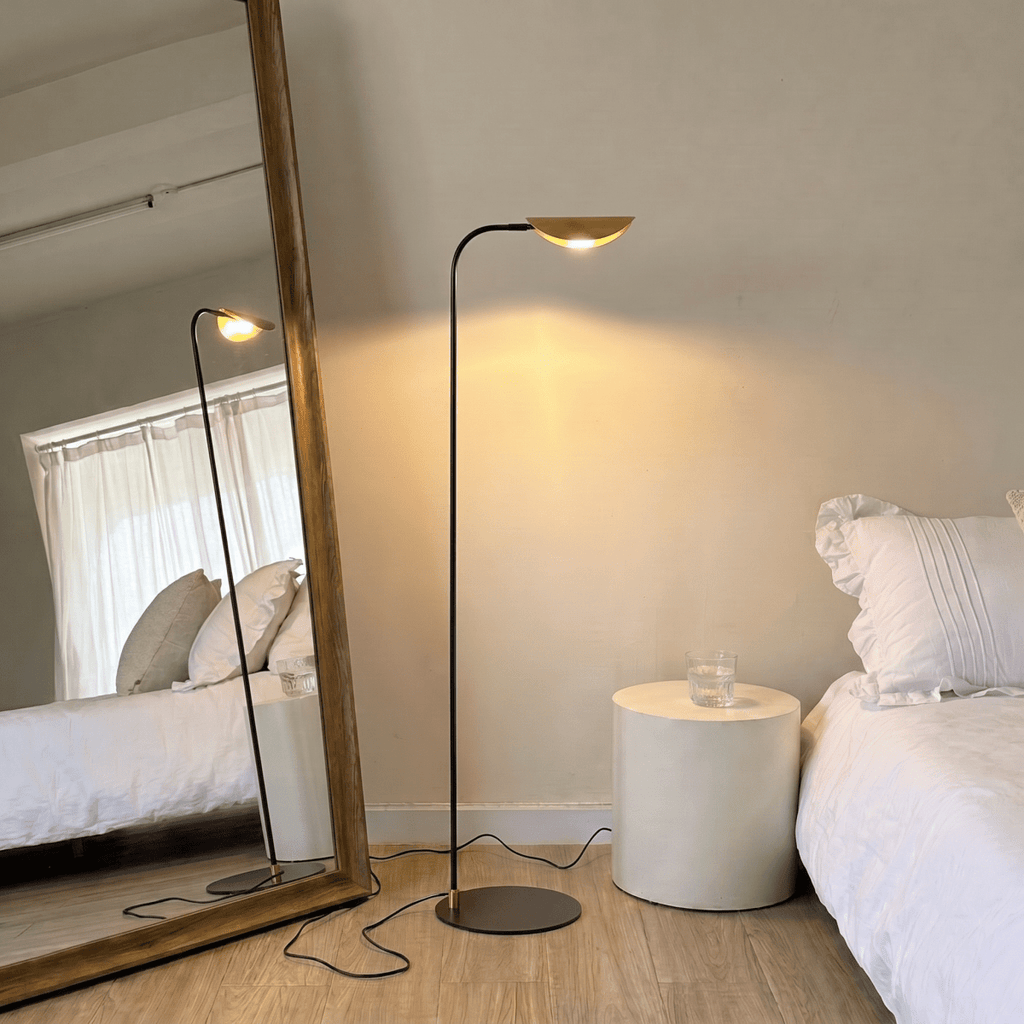 A brass-and-black Clérachic floor lamp lights a bedroom corner beside a leaning mirror and a white bed.