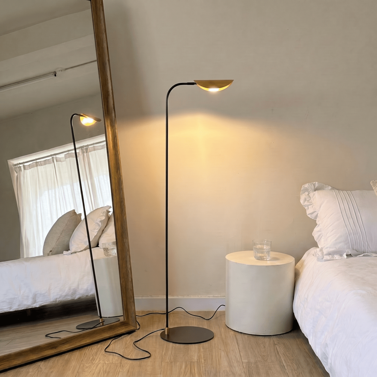 A brass-and-black Clérachic floor lamp lights a bedroom corner beside a leaning mirror and a white bed.