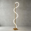 Bright white light glows along the looping silicone strip of this Clérachic floor lamp, on a brass base against a grey concrete wall.