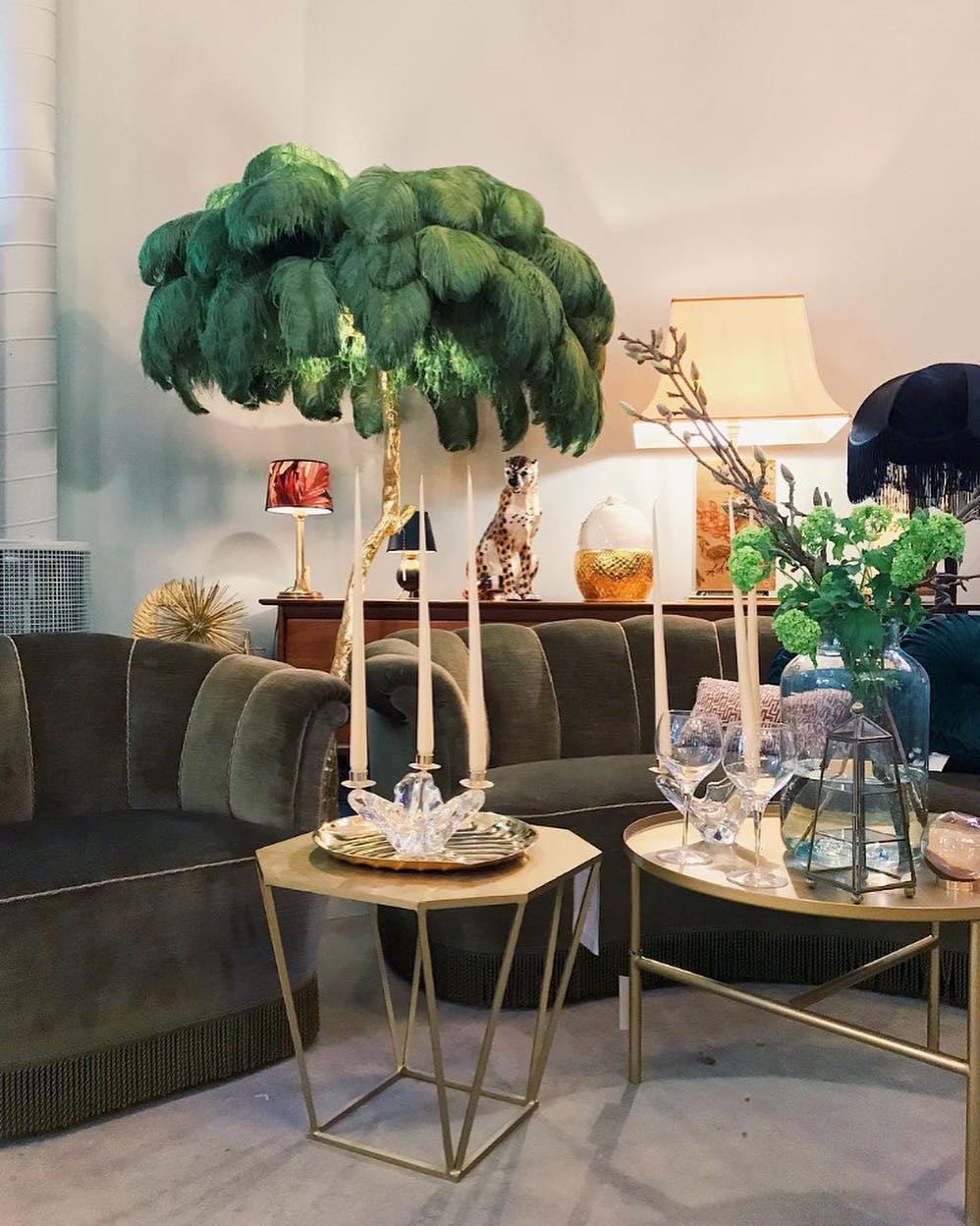 Deep green plumes over a Clérachic floor lamp in a lounge with olive velvet sofas, brass hexagonal tables and candles.
