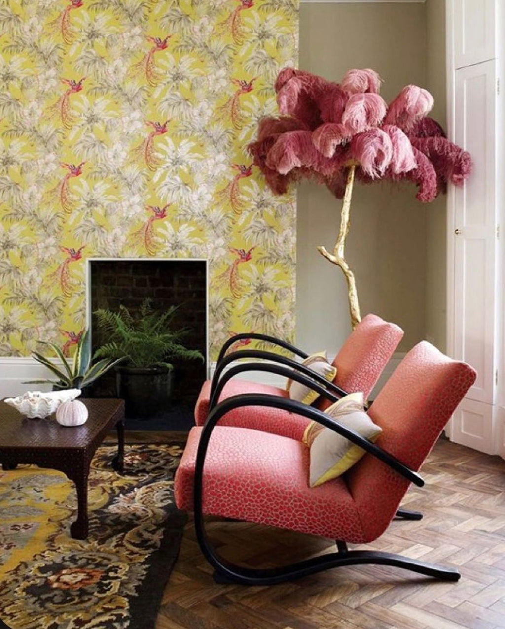 Dusky rose plumes on a Clérachic floor lamp against yellow botanical wallpaper, beside coral armchairs and a patterned rug.