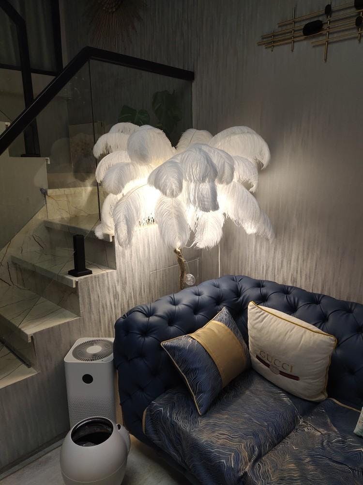 Glowing white plumes on a lit Clérachic floor lamp beside a navy tufted sofa, at the foot of a glass-balustrade staircase.