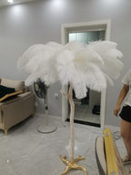 White feathers on a Clérachic floor lamp in a grey room, with its gold base on a marble floor and a boxed carton nearby.