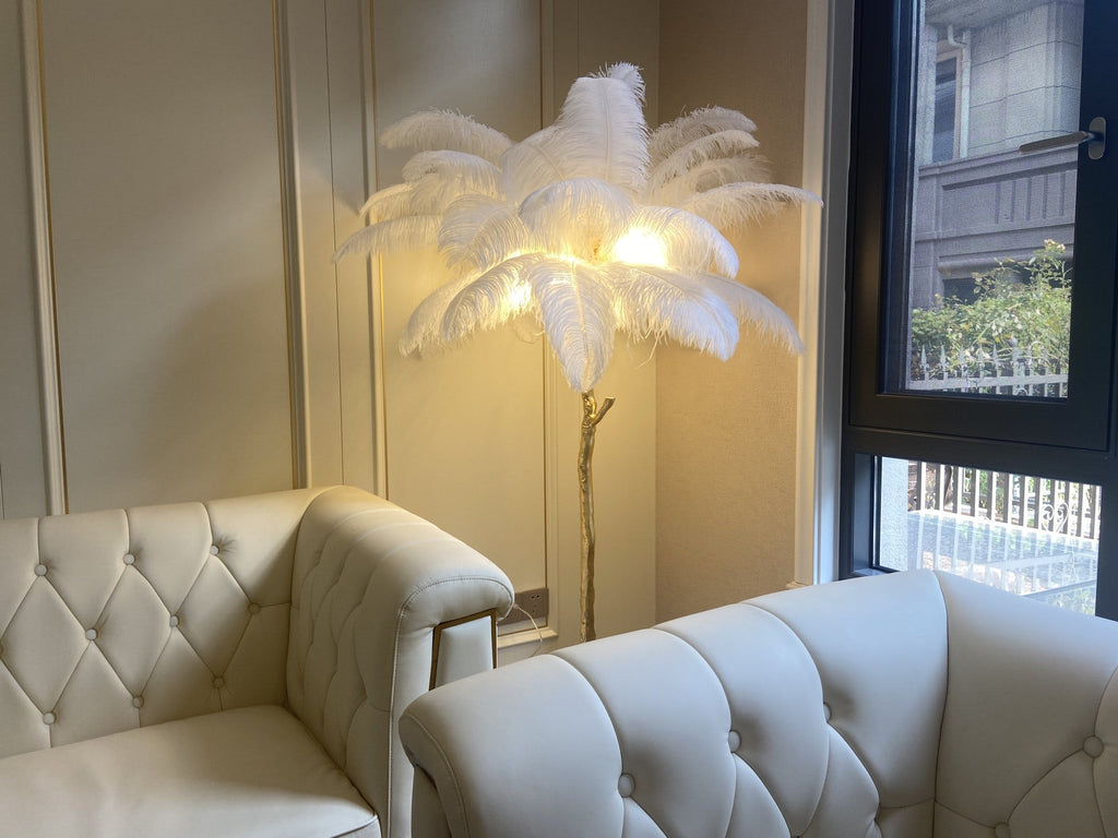 A lit Clérachic floor lamp with white plumes glows warmly in a corner between two cream tufted leather sofas and a window.