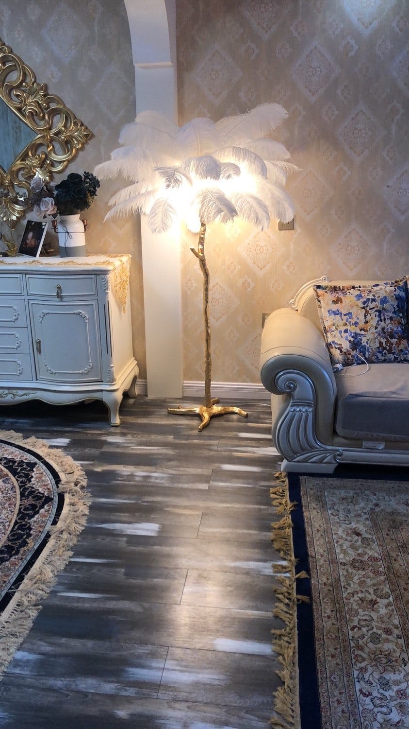 A lit white-feathered Clérachic floor lamp glows in an ornate sitting room, with a dark parquet floor and a blue sofa.