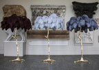Three Clérachic floor lamps stand in a row in a gallery space, with plum, pale blue and dark blue-grey plumes on gold stems.