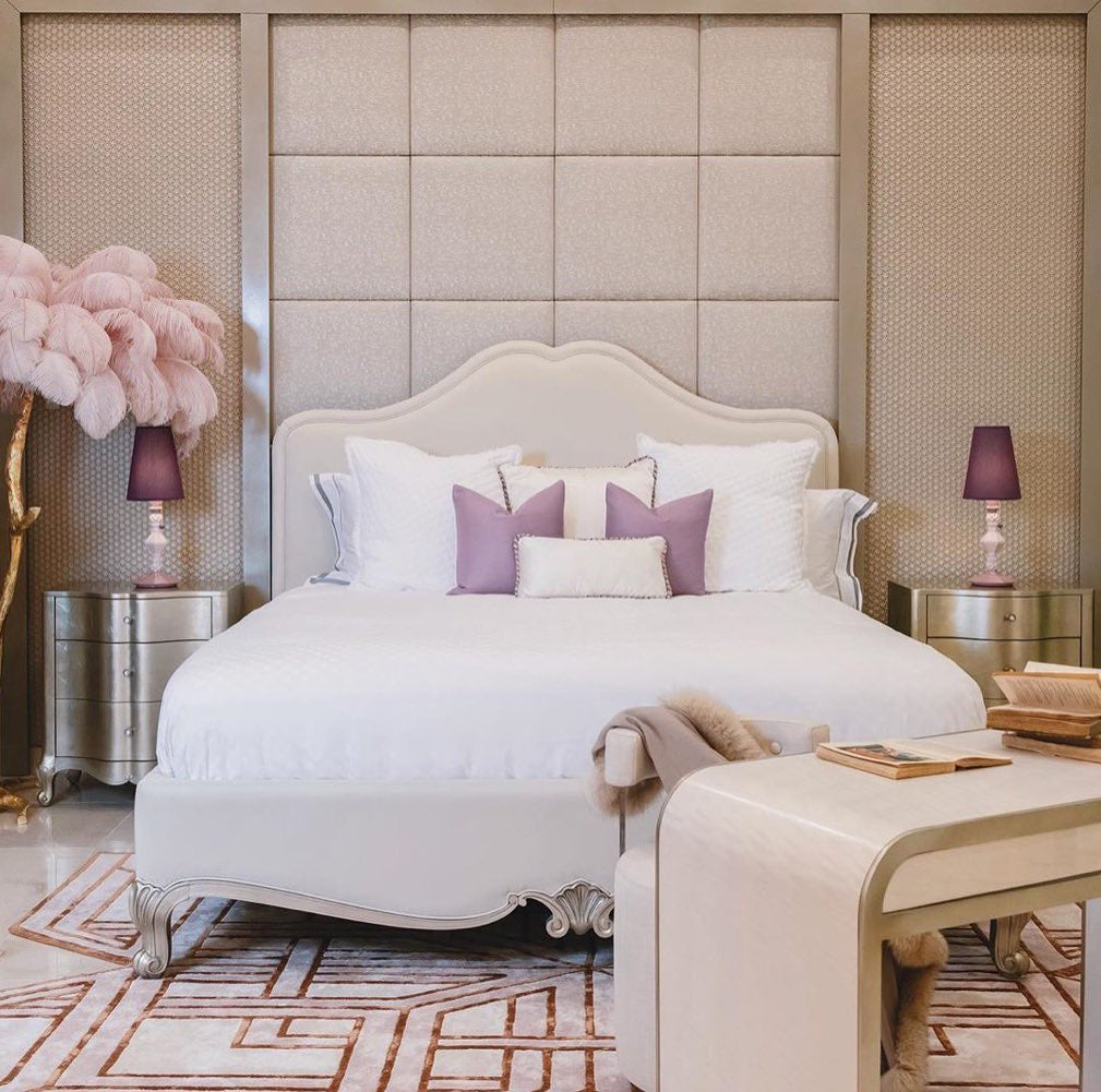 Pink plumes of a Clérachic floor lamp at the left edge of a cream bedroom, beside a white bed and lilac lamps.