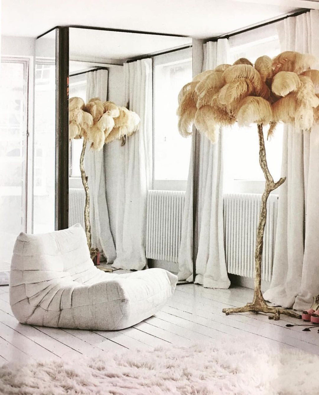 Two Clérachic floor lamps with sand-coloured plumes beside a white lounge seat, in front of long white curtains.