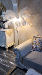White plumes lit warmly on a Clérachic floor lamp in an ornate sitting room, beside a pale cabinet and a blue sofa.