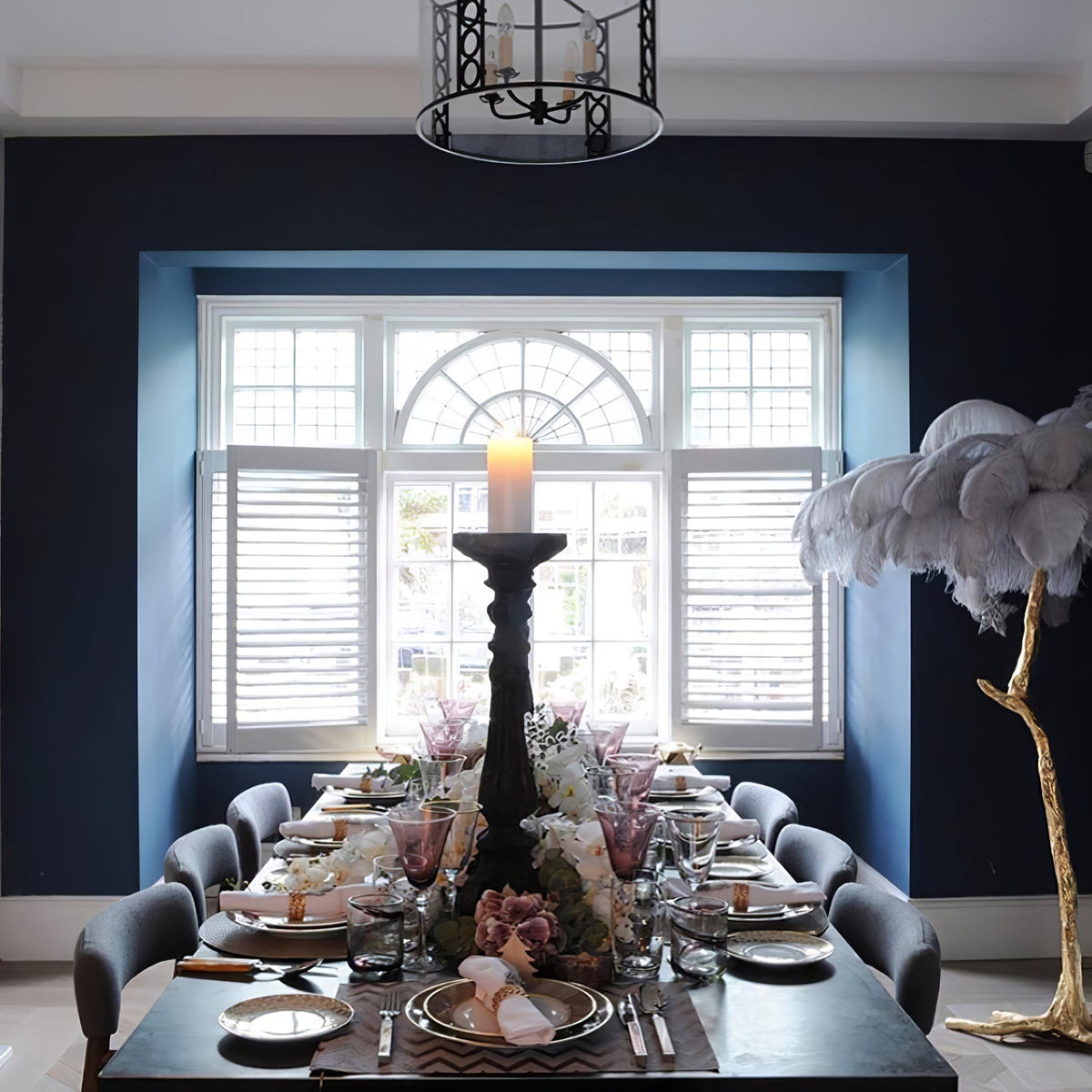 White-grey plumes on a Clérachic floor lamp at the edge of a navy dining room, behind a set table with a tall candle.