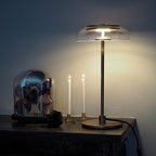 A lit Clérachic table lamp with a dark stem and clear glass dome on a dark ledge, beside two white candles and a glass cloche.