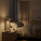 A Clérachic table lamp glowing on a bedside table in a dim bedroom, beside a cup and a stack of books, with a rumpled bed behind.